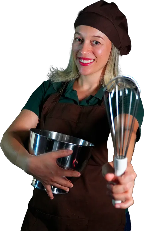 Dani Brasileiro de touca e avental marrons, sorrindo, com uma tigela de inox em um braço e um batedor de arame estendido para a frente